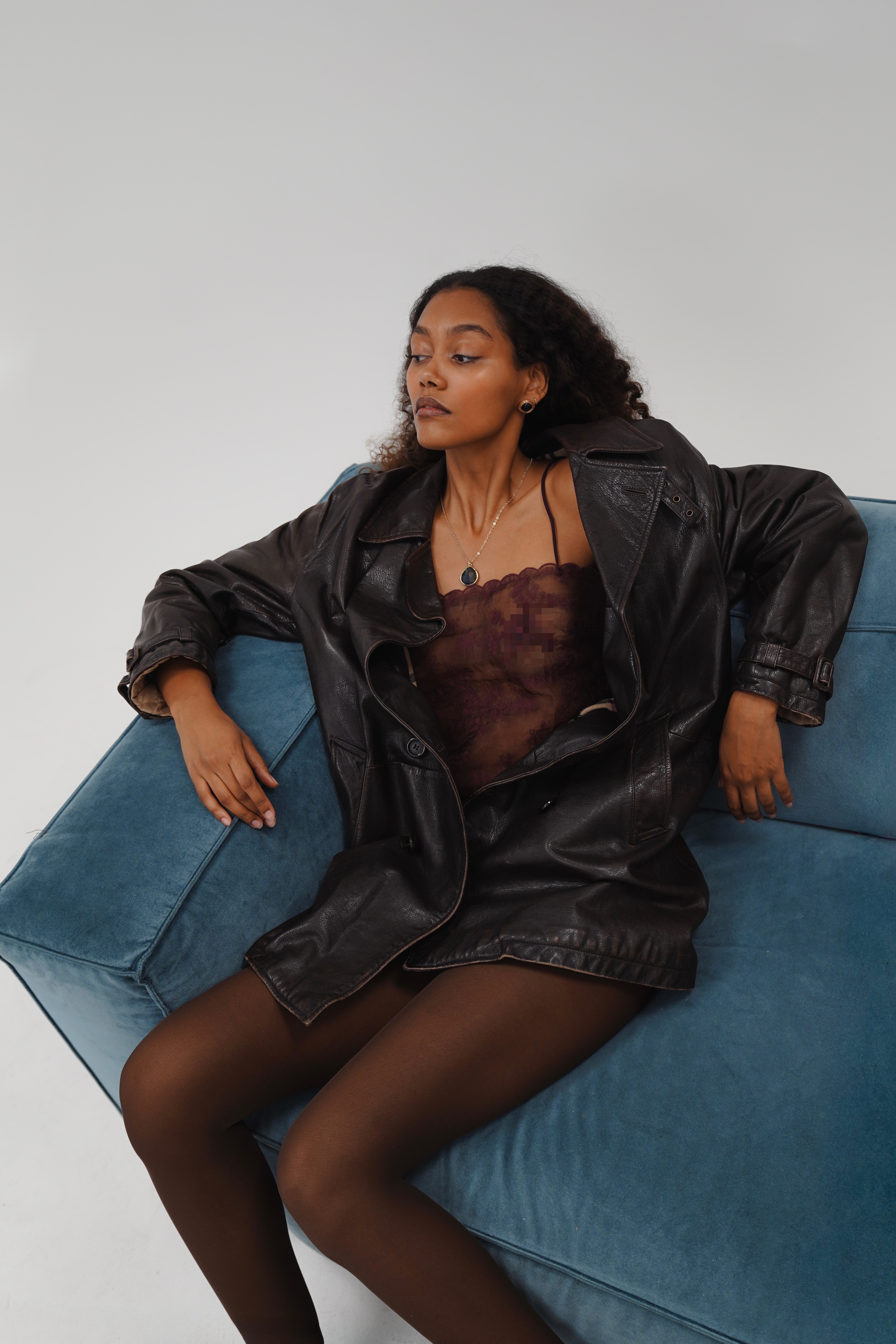 Woman with curly hair in black leather jacket over burgundy lace top, sitting on blue velvet armchair