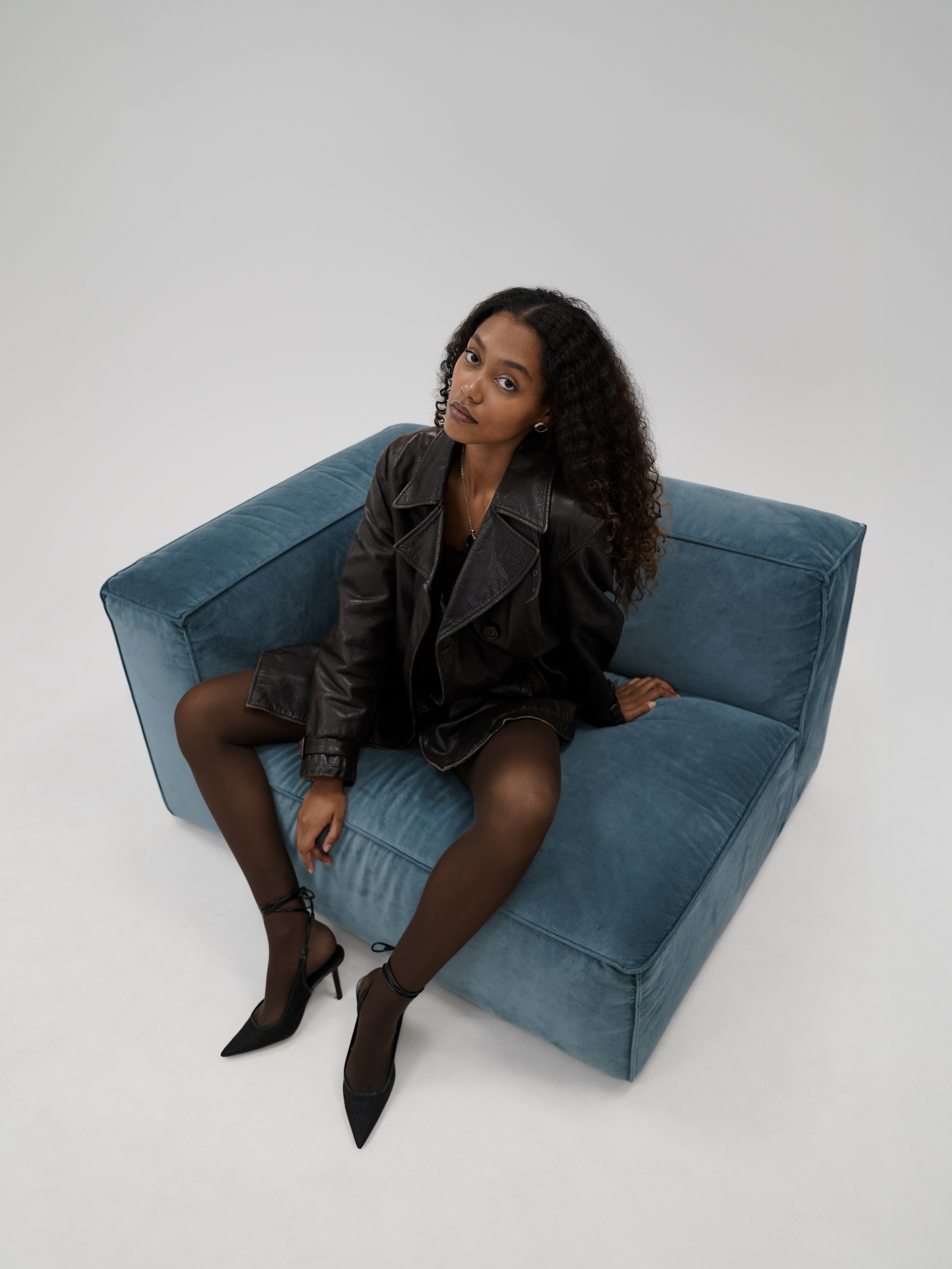 Woman with curly hair in black leather jacket sitting on blue velvet armchair
