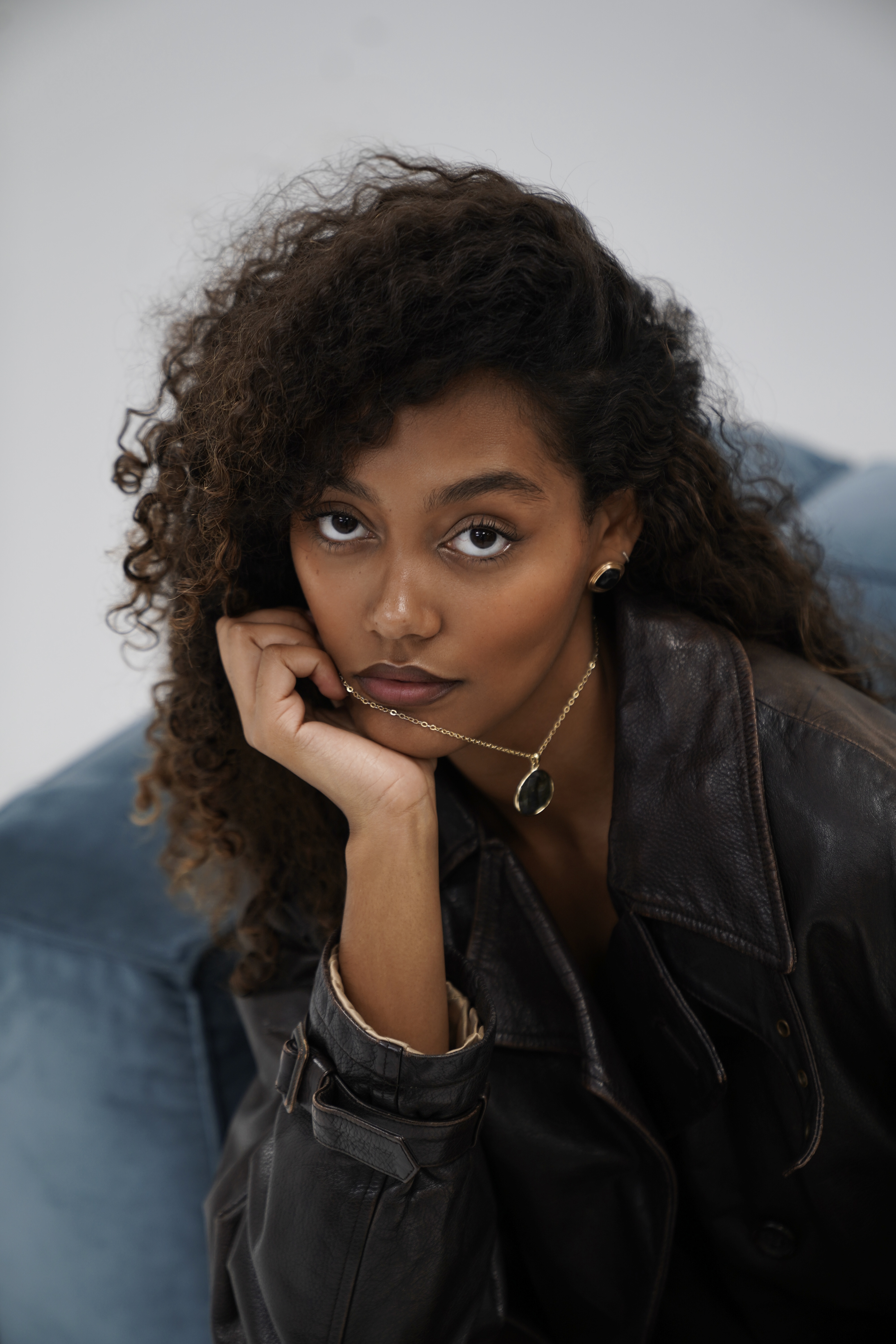 Woman with curly hair in black leather jacket and gold necklace, resting chin on hand